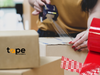 woman showing application of the "OPENED VOID" tape onto some cardoboard shipping boxes, tape providers logo on a box