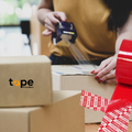 woman showing application of the  "OPENED VOID" tape onto some cardoboard shipping boxes, tape providers logo on a box